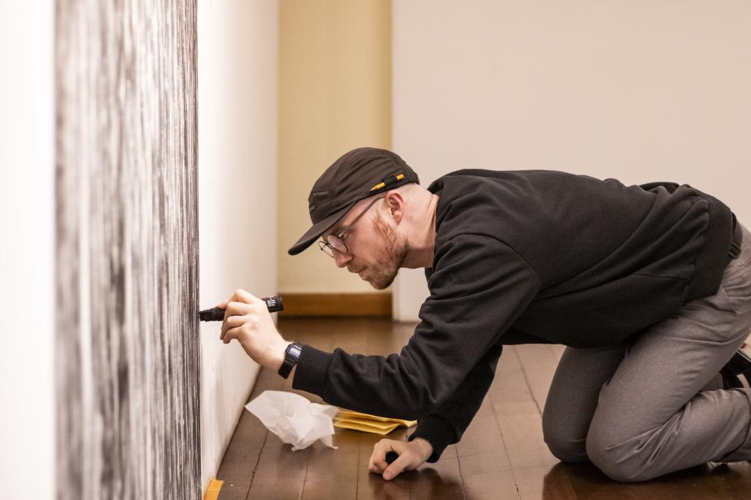 Um homem traçando uma sequência de linhas verticais pretas de 171cm de altura sobre uma parede.