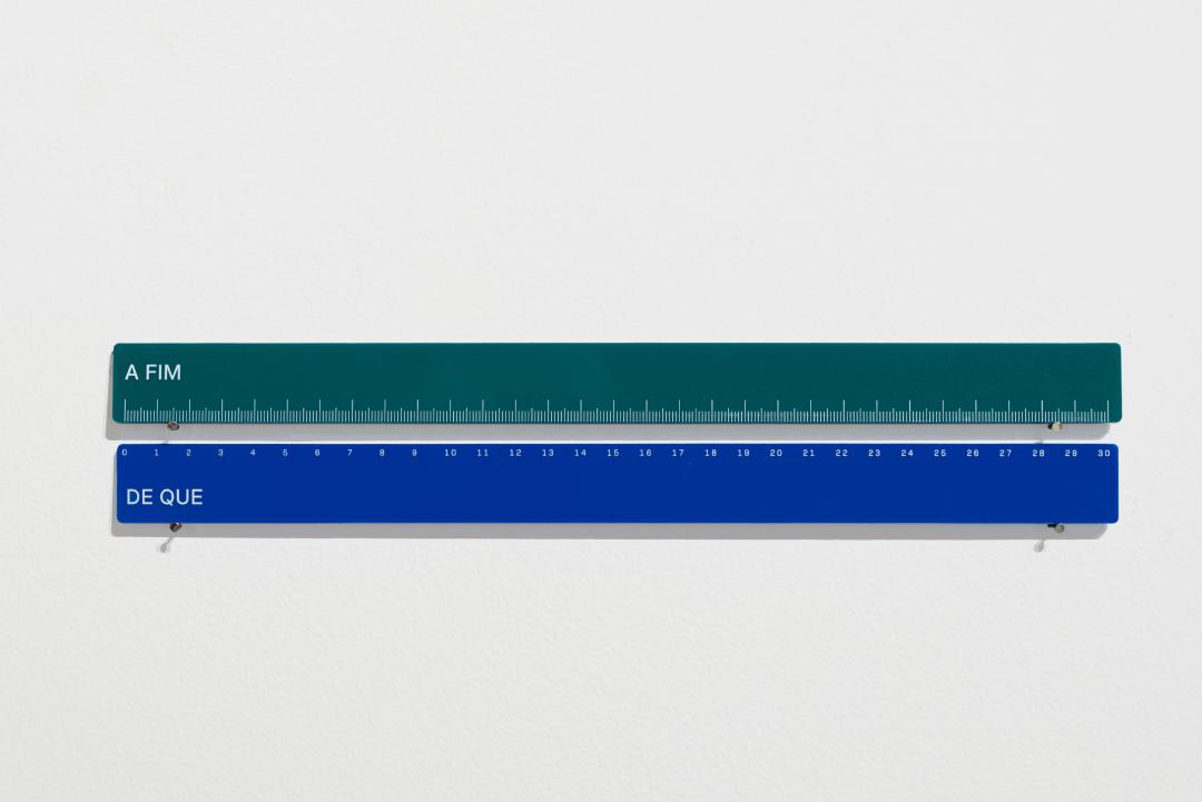 Duas réguas de 30 centímetros verde e azul, uma acima da outra. Na primeira somente os números dos centímetos e na segunda somente as marcações dos centímetros. Na de cima está escrito: &ldquo;A fim&rdquo;. E na de baixo: &ldquo;De que&rdquo;.