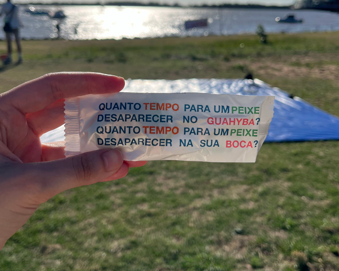 Uma mão segura uma embalagem onde se lê: &ldquo;Quanto tempo para um peixe desaparecer no Guahyba? Quanto tempo para um peixe desaparecer na sua boca?&rdquo;.