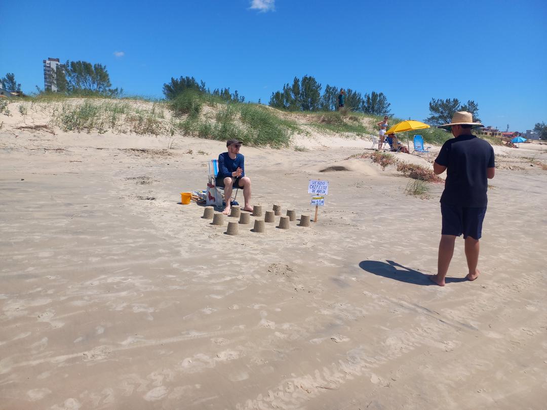 Na faixa de areia de uma praia um homem está sentado em uma cadeira de praia com 12 castelos de areia em sua frente, ao lado dos castelos uma placa com os dizeres &ldquo;Vendo castelos de areia: R$10,00&rdquo;, outro homem está parado olhando os castelos e a placa.